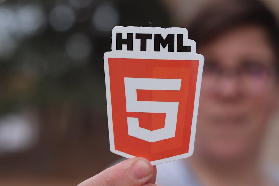 A person holds a sticker with the HTML5 logo, which features a white shield shape with an orange background and a bold white number 5 in the center. The person's fingers are visible grasping the stick