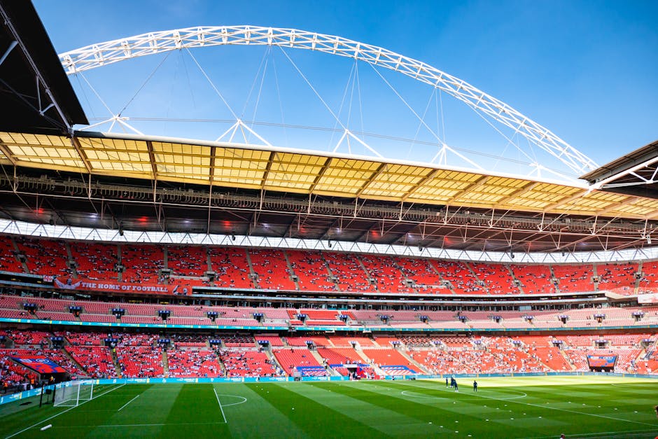 A large, modern stadium with tiered seating in shades of red and orange, arranged in multiple levels around a central grass pitch. The stadium's open design features a prominent white steel truss roof